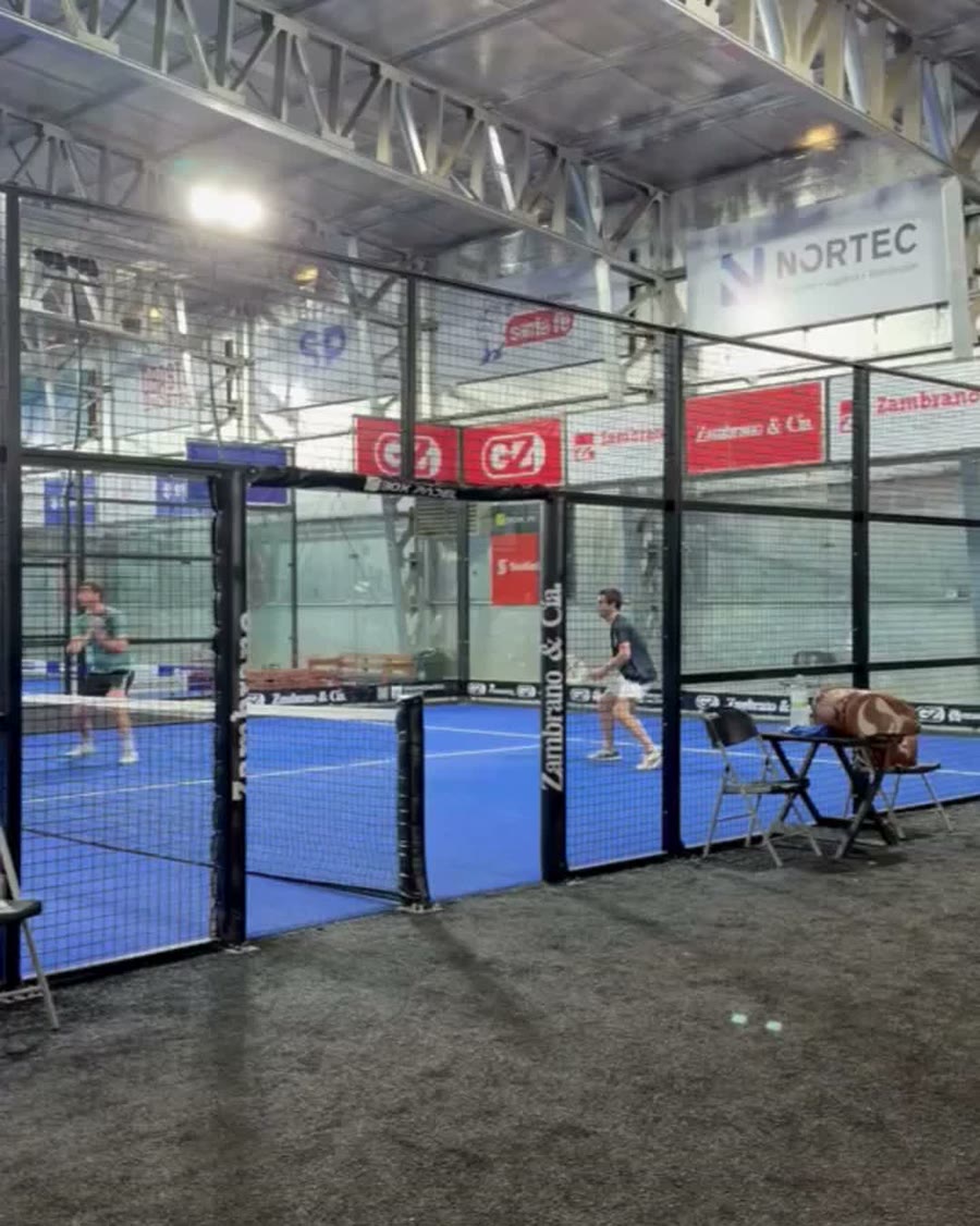 Canchas de cristal y césped azul bajo el techo del galpón, con jugadores en pleno partido.